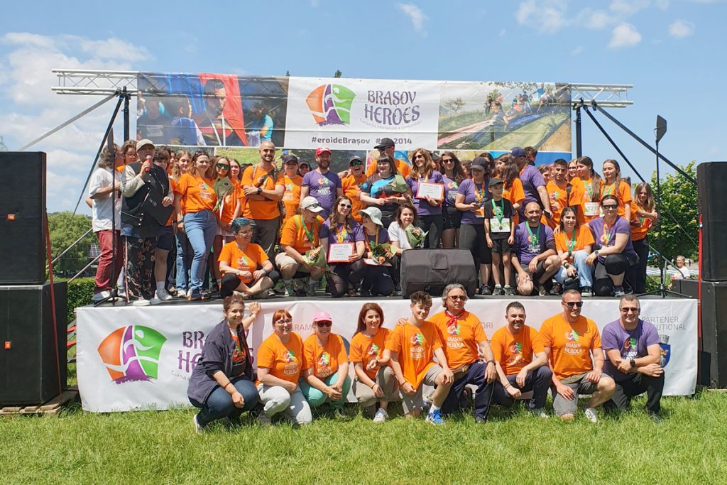 Voluntarii și organizatorii Brașov Heroes 2025, fotografiați pe scenă în tricouri colorate, alături de susținători