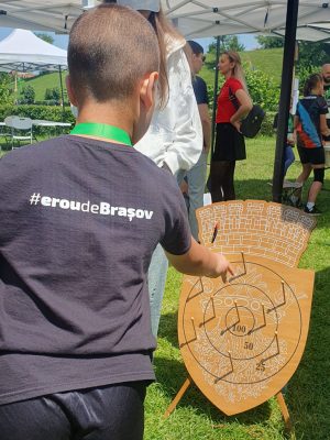 Copil cu tricou insccriptionat Erou de Brasov aruncă cu inele din cauciuc la jocul din lemn de Aruncare la țintă cu Stema Brașov imprimată pe țintă