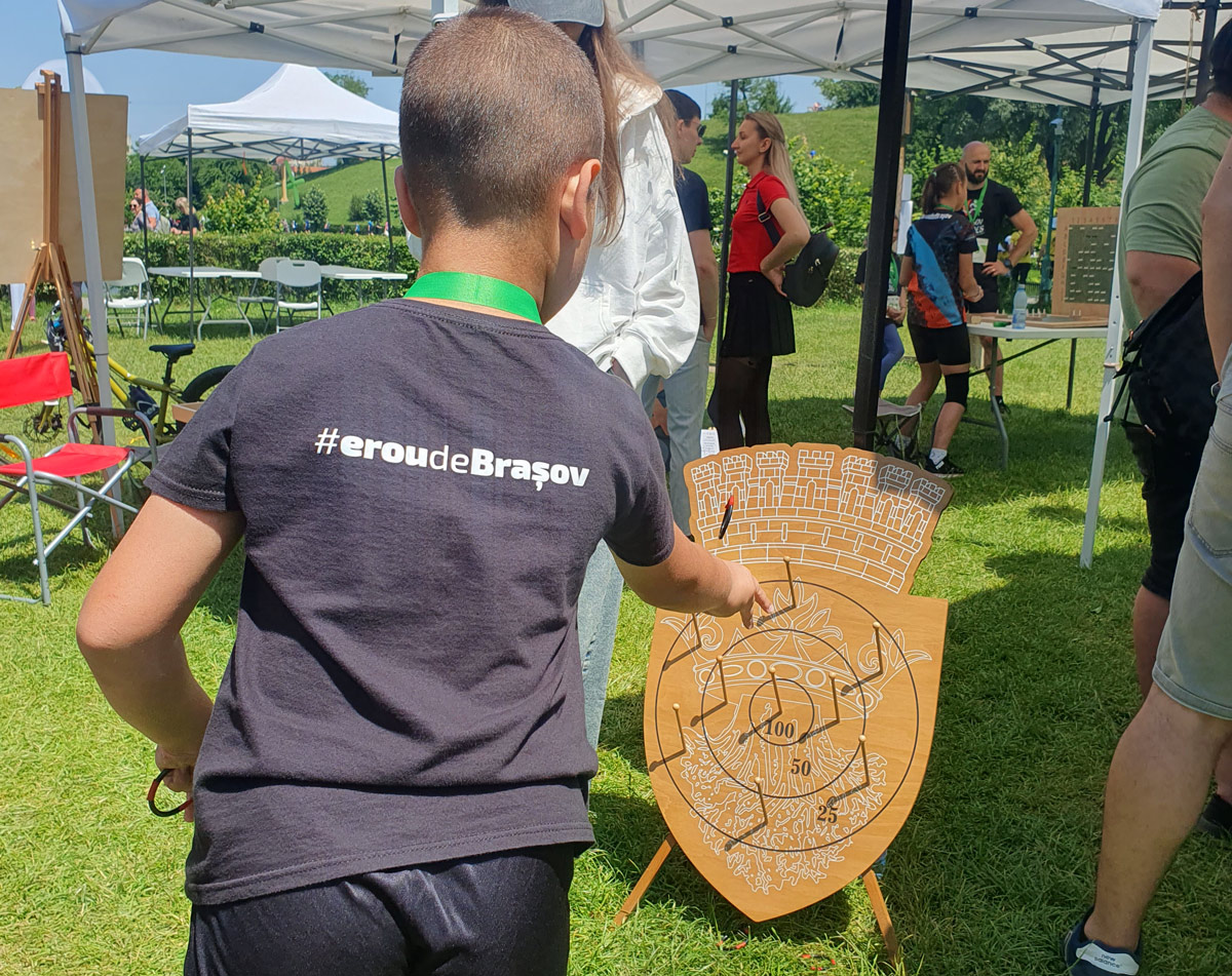 Copil cu tricou insccriptionat Erou de Brasov aruncă cu inele din cauciuc la jocul din lemn de Aruncare la țintă cu Stema Brașov imprimată pe țintă