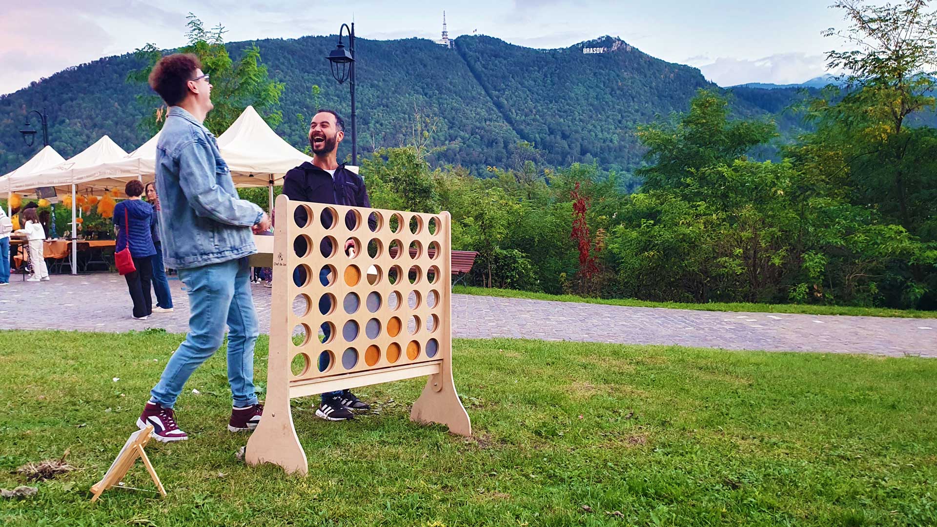 2 youg men lauthing and playng Giant Connect Four at Brașov citadel woith mountain Tampa in the back