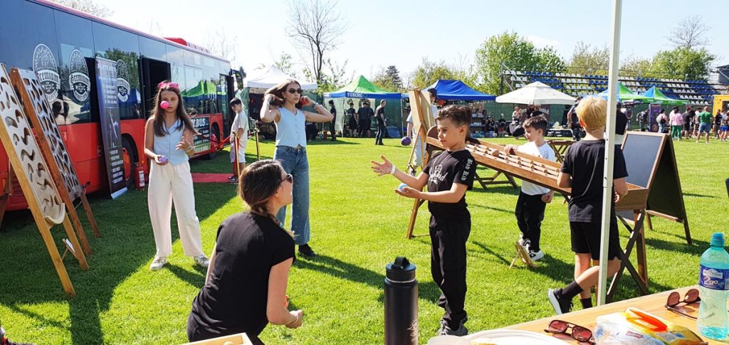 Animatoare Chef de Joc predând jongleria unui grup de copii la festival, cu jocuri din lemn în fundal