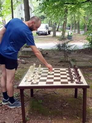 Bărbat jucând șah gigant din lemn în aer liber, tablă de 105×105 cm la înălțime confortabilă, picioare vizibile, cadru natural verde